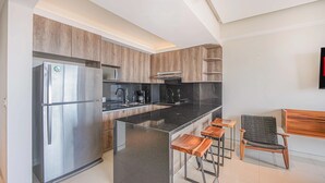A modern kitchen with a stainless steel refrigerator, black countertops, wooden cabinets, and a bar area with stools. - Torre Ku, Ocean Views Near the Beach (Puerto Vallarta)