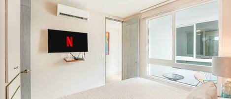 A bedroom with a bed, a mounted TV displaying the Netflix logo, a window, and a door leading to another room.