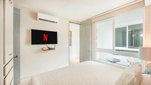 Ein Schlafzimmer mit einem Bett, einem an der Wand befestigten Fernseher mit dem Netflix-Logo, einem Fenster und einer Tür zu einem weiteren Raum.