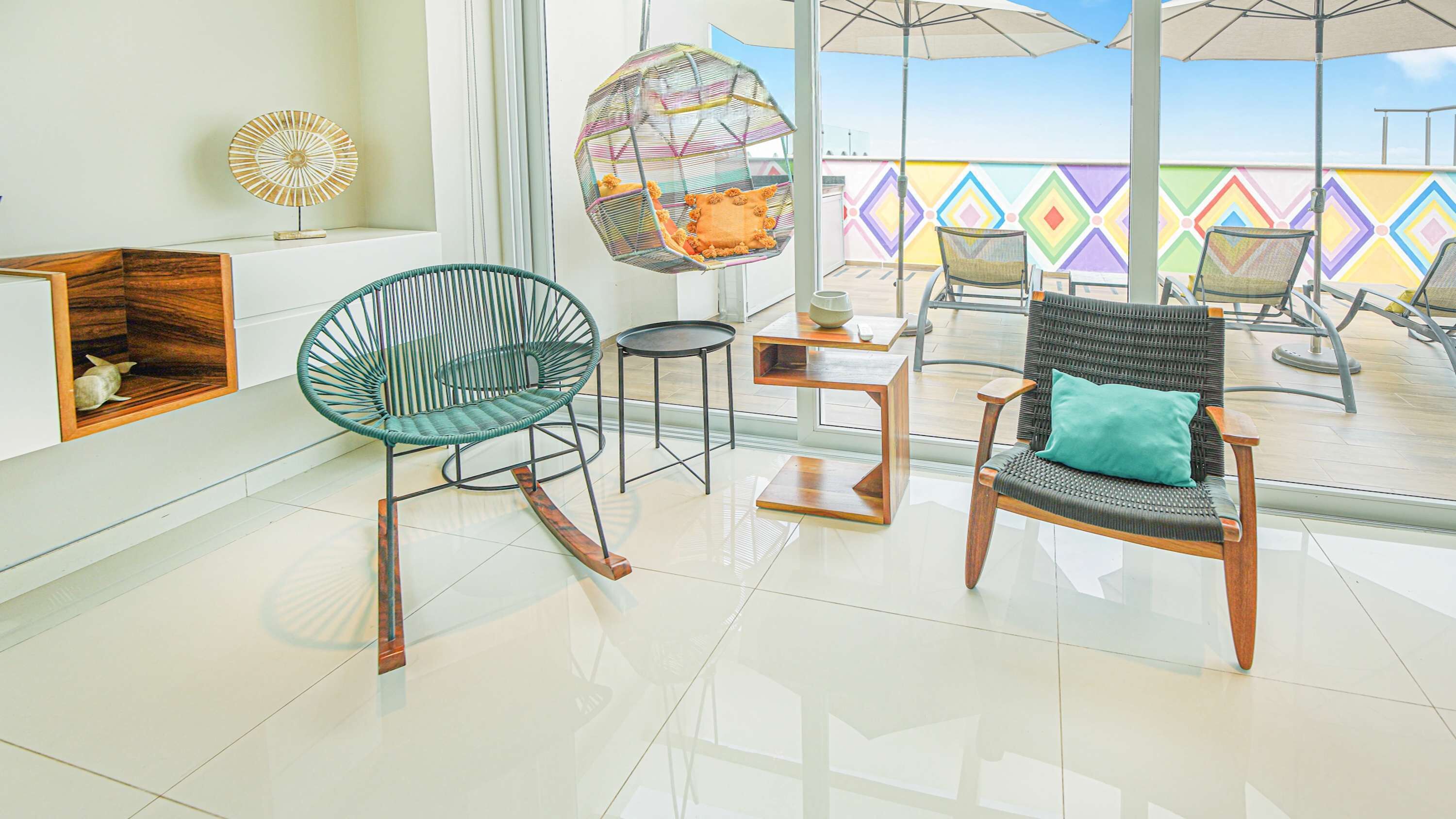 A modern interior with a glass floor, a green wicker chair, a black wicker chair with a teal cushion, and a hanging chair with a colorful cushion.