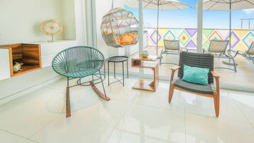 A modern interior with a glass floor, a green wicker chair, a black wicker chair with a teal cushion, and a hanging chair with a colorful cushion.