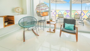 A modern interior with a glass floor, a green wicker chair, a black wicker chair with a teal cushion, and a hanging chair with a colorful cushion. - Torre Ku · Rooftop Pool with Ocean Views (Puerto Vallarta)