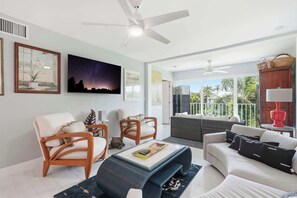 Ein modernes Wohnzimmer mit Deckenventilator, Couchtisch, zwei Sesseln, einer Couch und Blick ins Freie durch große Fenster.
