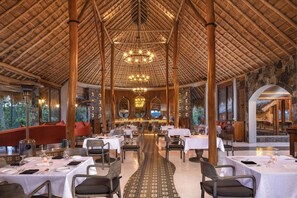 An indoor dining area with tables and chairs, a central walkway, and a high ceiling with a wooden structure. - 98 Acres Resort and Spa (Ella)