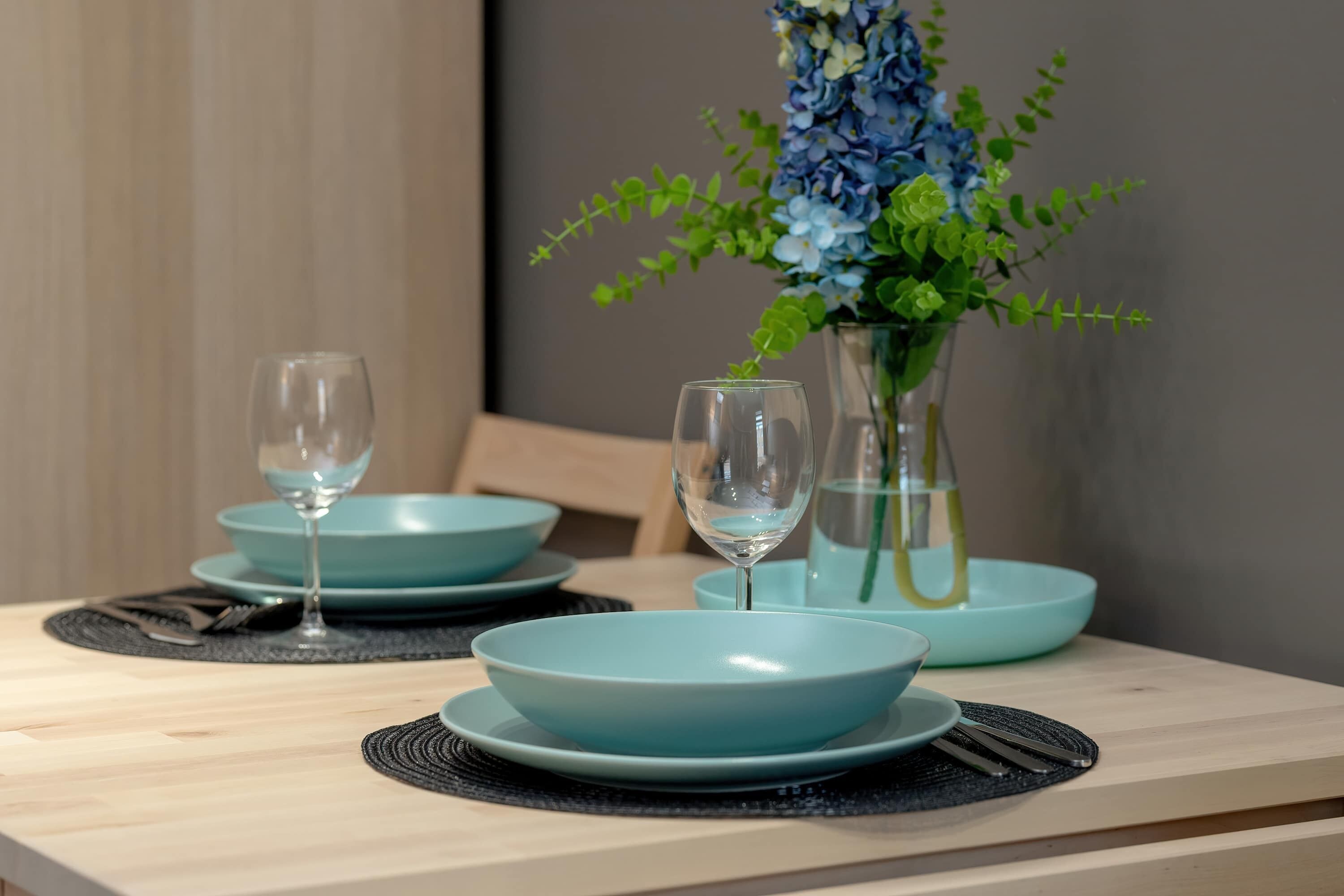 A dining table with two blue plates, a vase with flowers, and two wine glasses.