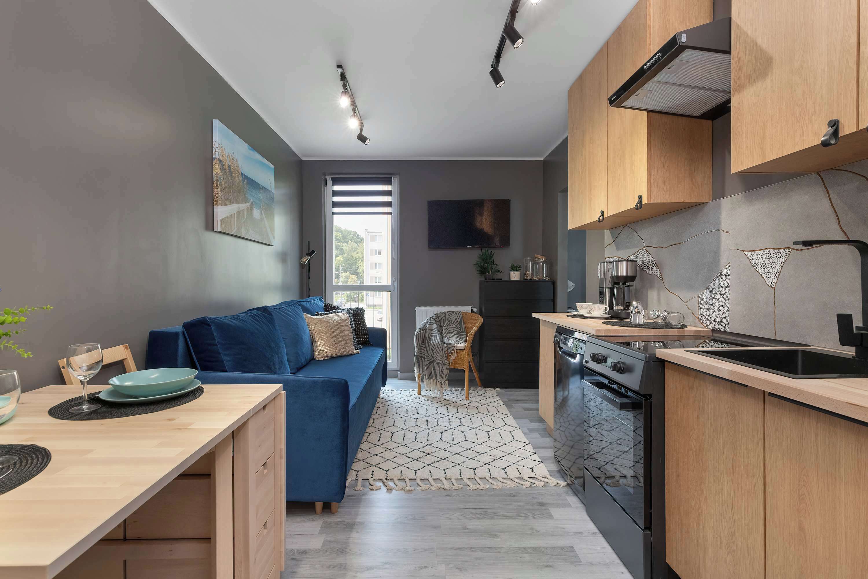 A modern kitchen with a built-in oven, a dining area with a blue sofa, and a large window providing natural light.