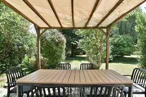 Eine überdachte Terrasse im Freien mit einem Holztisch und Stühlen, umgeben von üppigem Grün.