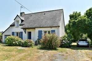 Une maison à deux étages, avec un toit en pente, une cheminée bien visible et une voiture garée dans l’allée.