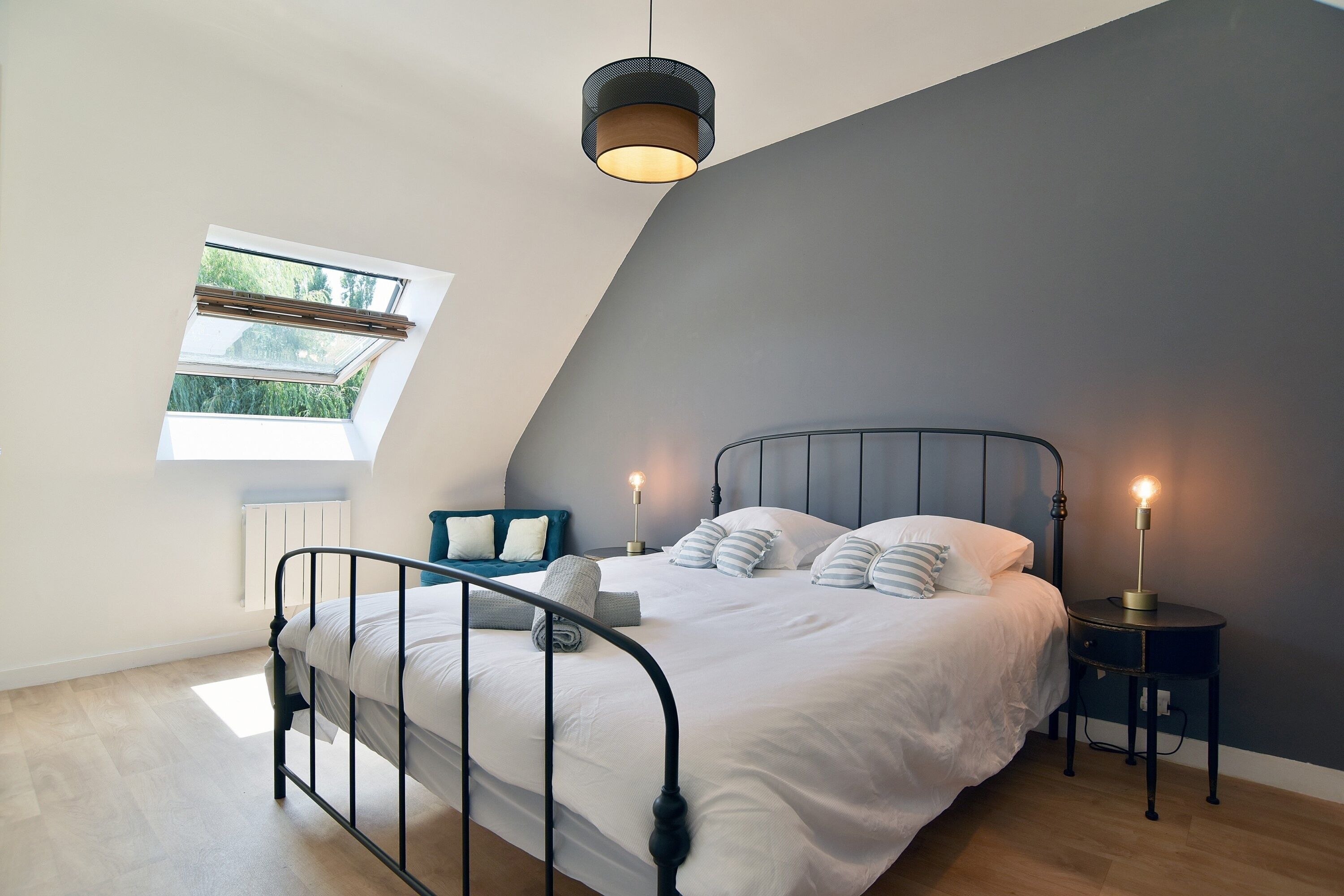 A bedroom with a sloped ceiling, a bed with white bedding, two bedside lamps, and a window with a view of greenery.