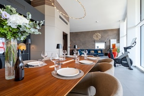 Ein moderner Essbereich mit einem langen Holztisch, der für eine Mahlzeit gedeckt ist, ein Fitnessbereich mit einem Ergometer und ein Wohnraum mit einem blauen Sofa und einem großen Fenster.