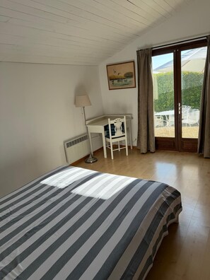 Een kamer met een bed, een bureau, een stoel, een radiator en uitzicht op groen via een deur.