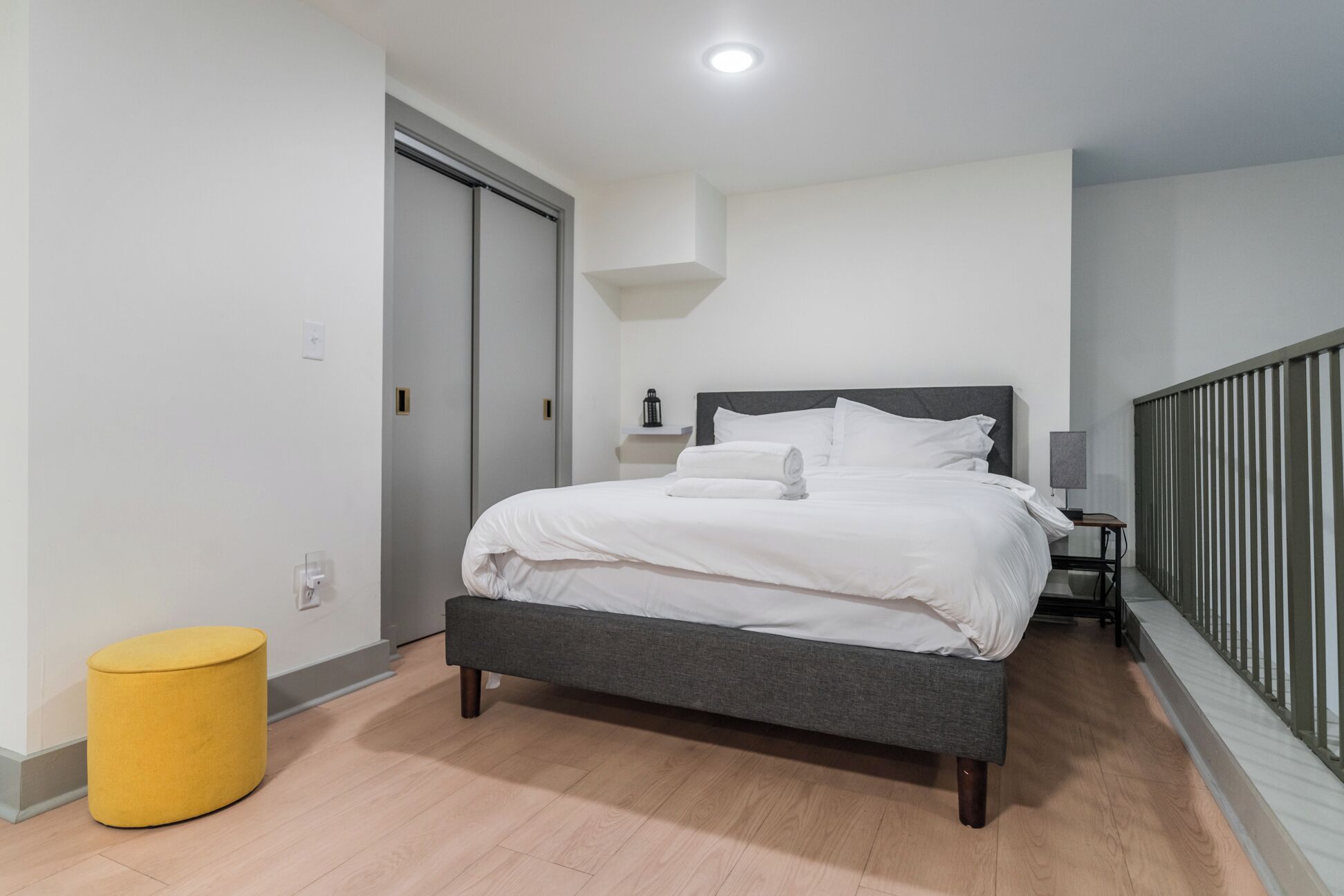 Une chambre à coucher moderne avec un lit, une table de chevet, une lampe et un pouf jaune.