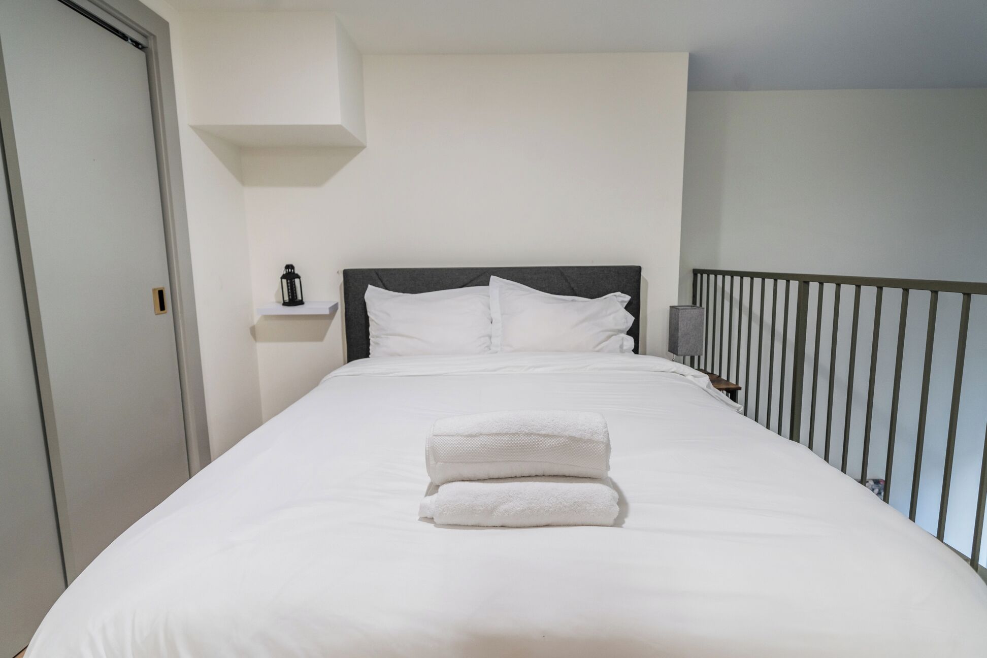 A neatly made bed with white linens and a folded towel, a bedside table with a lamp, and a view of the balcony railing.