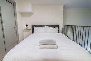 A neatly made bed with white linens and a folded towel, a bedside table with a lamp, and a view of the balcony railing. - Industrial Loft  - Nearby Parking Downtown - 2F (Louisville)
