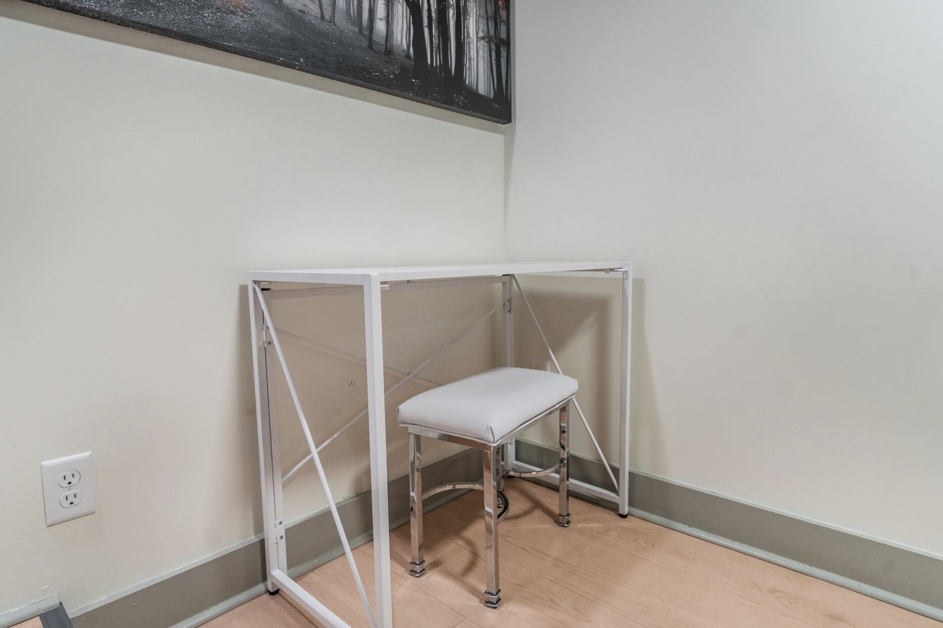 A room with a white metal desk and bench, a painting on the wall, and a light switch on the wall.