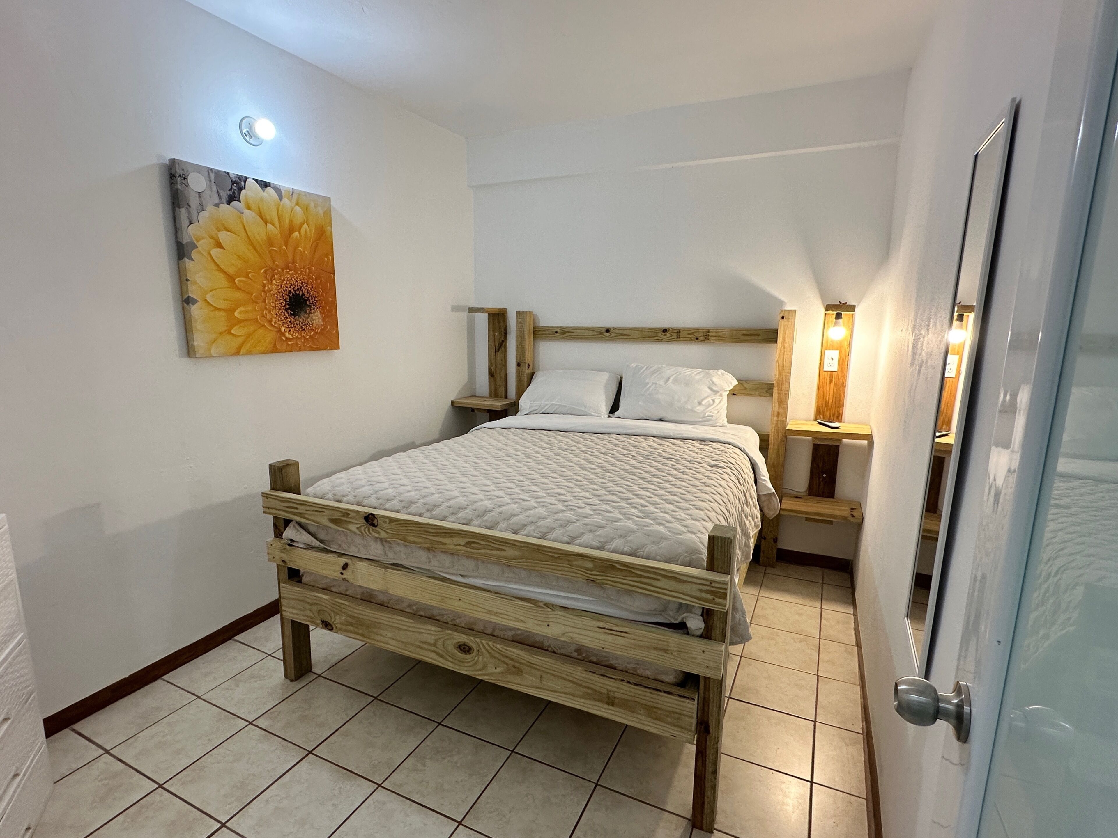 A single-bed room with a wooden frame, white bedding, a wall-mounted flower painting, bedside lamps, and a tiled floor.