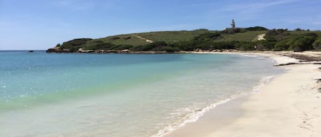 A beach with clear water, a sandy shore, and a hill with a lighthouse in the distance.