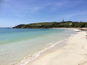 En strand med kristallklart turkost vatten, en sandstrand och en kulle med en fyr i fjärran.