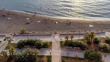 Una spiaggia con persone che nuotano e prendono il sole, un lungomare con panchine e palme.