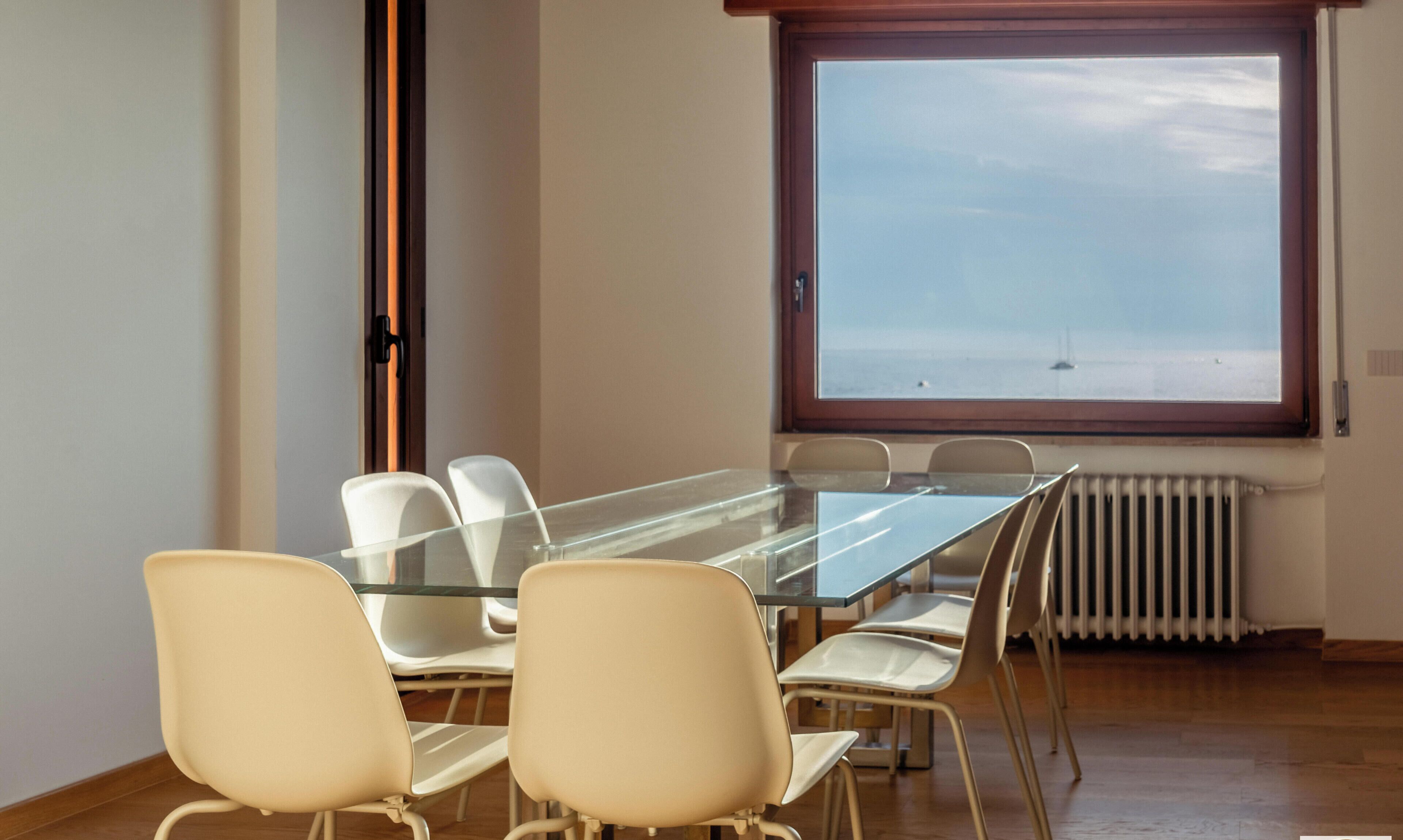 Ein Konferenzraum mit Glastisch und weißen Stühlen, ein Fenster mit Meerblick und ein Heizkörper darunter.