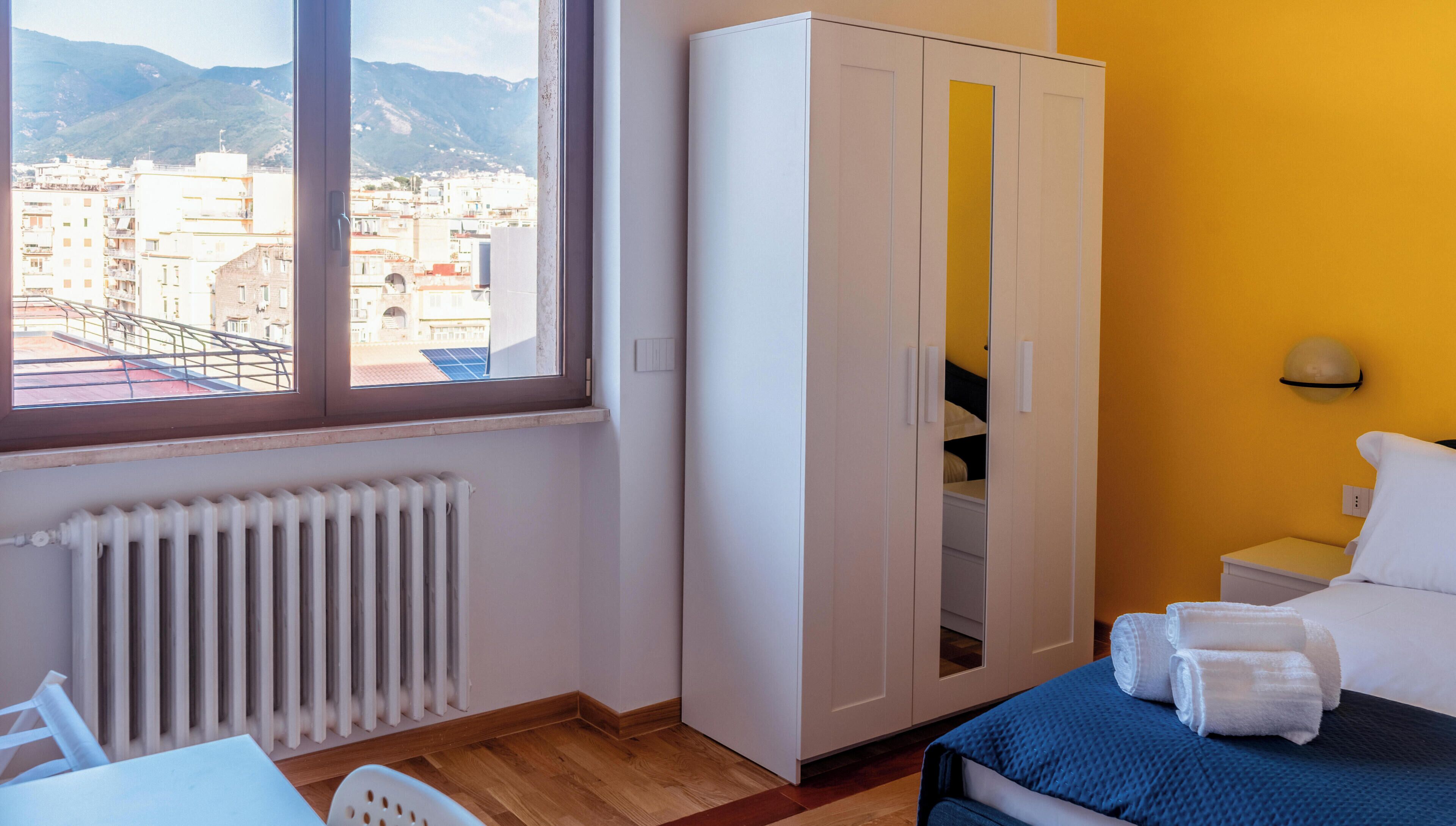 Une chambre à coucher comprenant un lit, une armoire, un radiateur, une fenêtre donnant sur des bâtiments et des montagnes, ainsi qu’une table de chevet avec des serviettes.