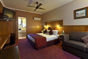 Een hotelkamer met een bed, een bank, een televisie, een plafondventilator en een badkamer zichtbaar door een geopende deur.