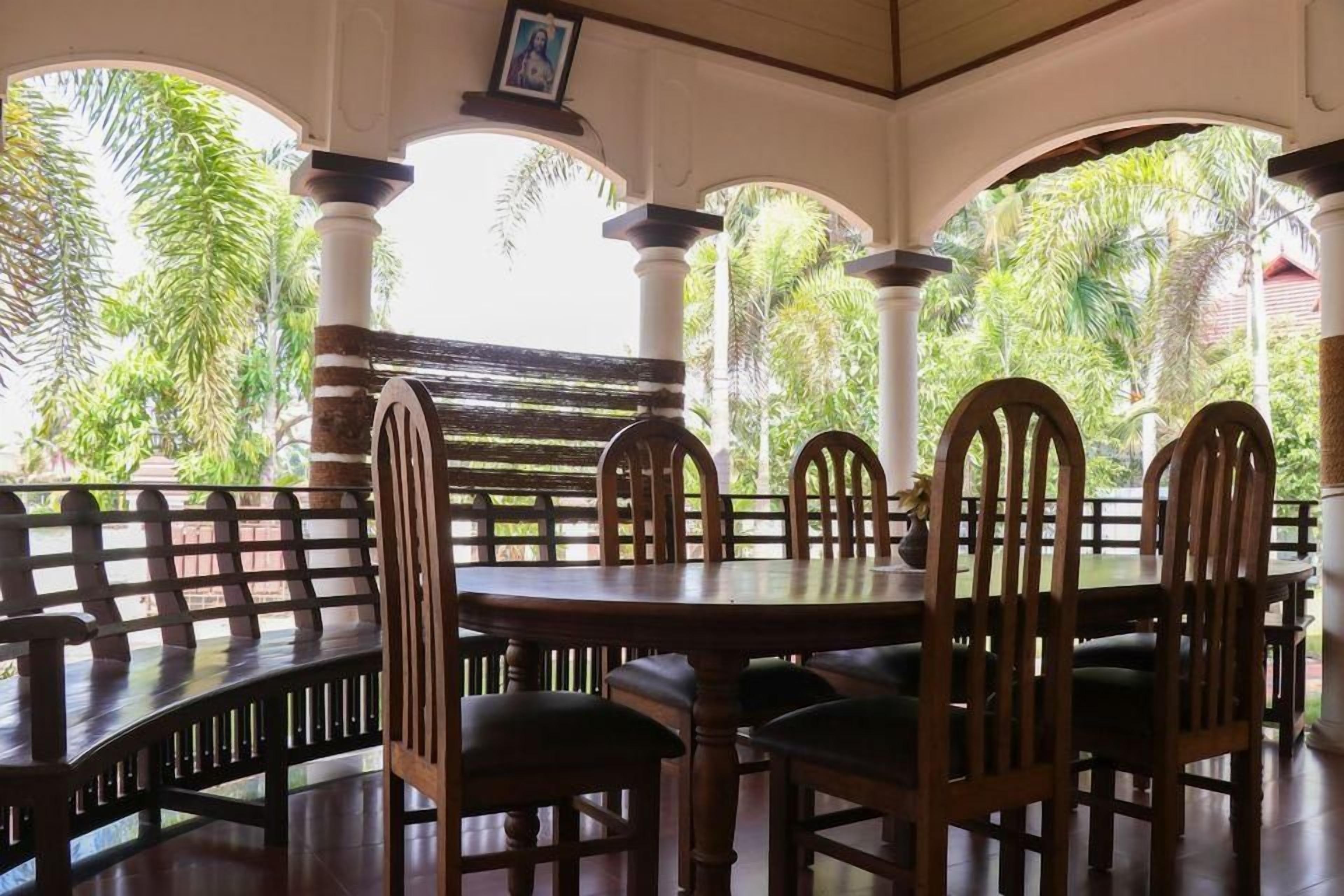 A covered outdoor seating area with a round table and chairs, surrounded by columns and a view of tropical foliage.