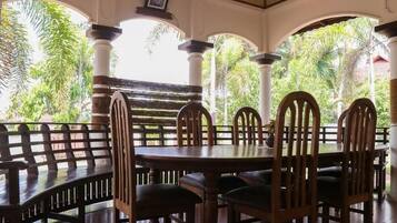 A covered outdoor seating area with a round table and chairs, surrounded by columns and a view of tropical foliage.