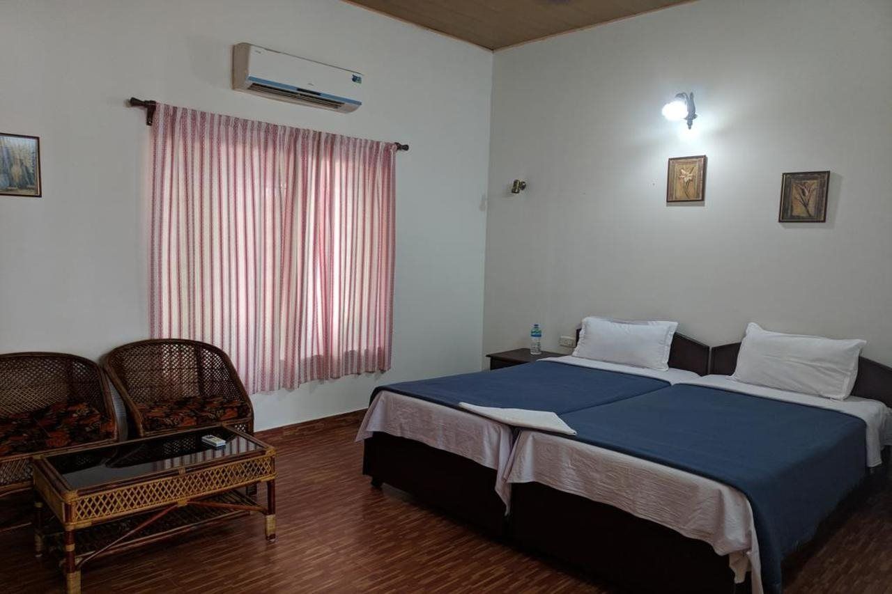 A room with a bed, two nightstands, a wall-mounted air conditioner, a window with curtains, and two wicker chairs.