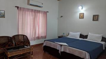 A room with a bed, two nightstands, a wall-mounted air conditioner, a window with curtains, and two wicker chairs.