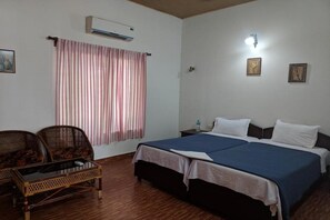 Una habitación con una cama, dos mesitas de noche, un aire acondicionado de pared, un ventanal con cortinas y dos sillones de mimbre.
