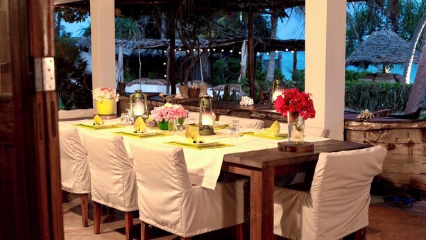 Un área de comedor al aire libre con una mesa preparada para cuatro personas, rodeada de sillas y decorada con flores y faroles.