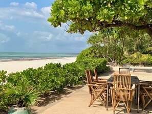 A beachside patio with wooden furniture, a view of the ocean, and lush greenery.