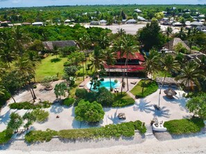 Vista aérea de un complejo turístico con una piscina central, rodeado de palmeras y una zona de playa arenosa.