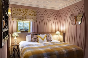 Quarto com cama, duas luminárias de cabeceira, janela com vista e painel decorativo na parede.