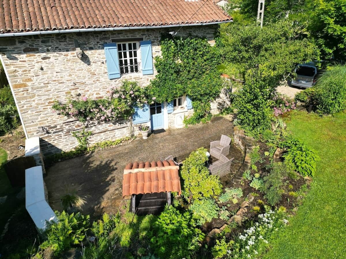 Uma casa de pedra com telhado de azulejos, um jardim com diversas plantas e um carro estacionado.