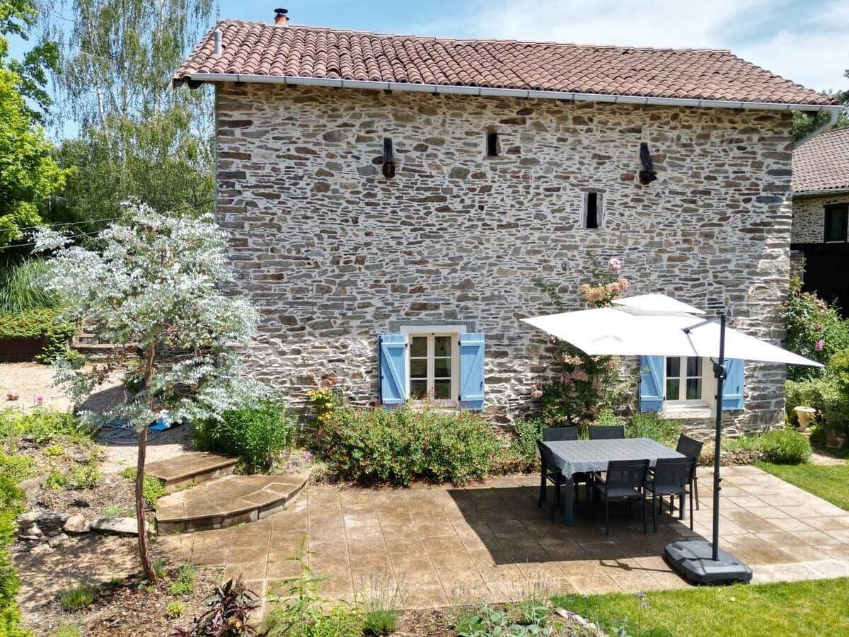 Une maison en pierre avec un toit en tuiles, une terrasse avec une table et des chaises, et un petit jardin agrémenté de plantes fleuries.