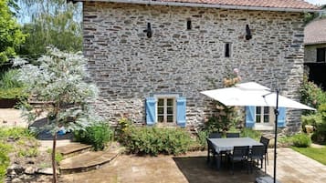 Une maison en pierre avec un toit en tuiles, une terrasse avec une table et des chaises, et un petit jardin agrémenté de plantes fleuries.