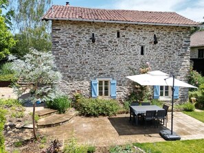 Ein Steinhaus mit Ziegeldach, eine Terrasse mit Tisch und Stühlen und ein kleiner Garten mit blühenden Pflanzen.