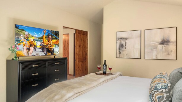 A hotel room with a bed, a television displaying a river rafting scene, a wooden door, and two abstract paintings on the wall. - Abode at The Timbers, minutes to skiing, hot tub (Ketchum)