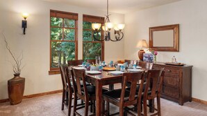 A dining area with a wooden table, chairs, a chandelier, and a view of trees through large windows. - Abode at The Timbers, minutes to skiing, hot tub (Ketchum)