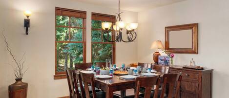 Un comedor con mesa de madera, sillas, una araña y vistas a árboles a través de amplios ventanales.