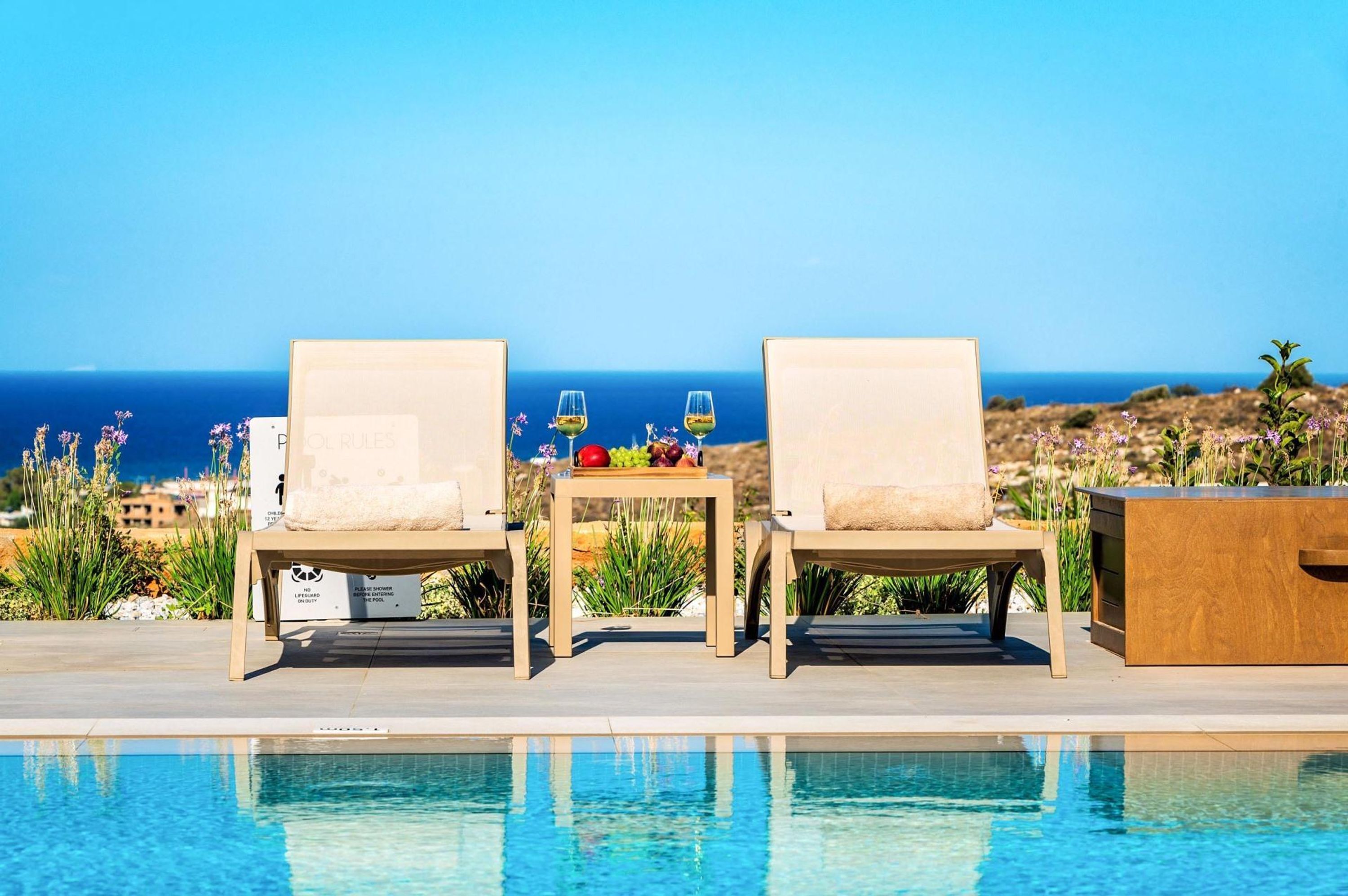 A poolside area with two lounge chairs facing a pool, a table with a fruit tray and glasses, and a view of the ocean.