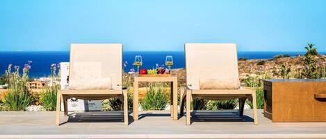 A poolside area with two lounge chairs facing a pool, a table with a fruit tray and glasses, and a view of the ocean.