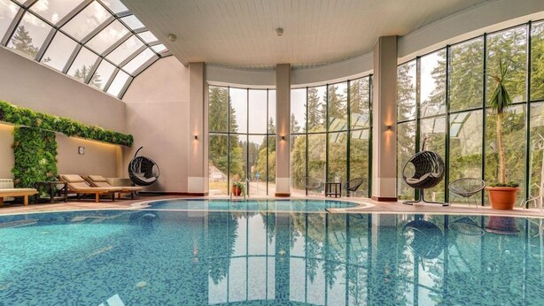 An indoor swimming pool with a glass ceiling, lounge chairs, and a view of trees. - Murgavets Hotel (Chepelare)