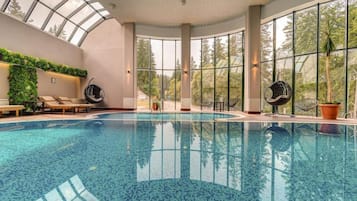 An indoor swimming pool with a glass ceiling, lounge chairs, and a view of trees.