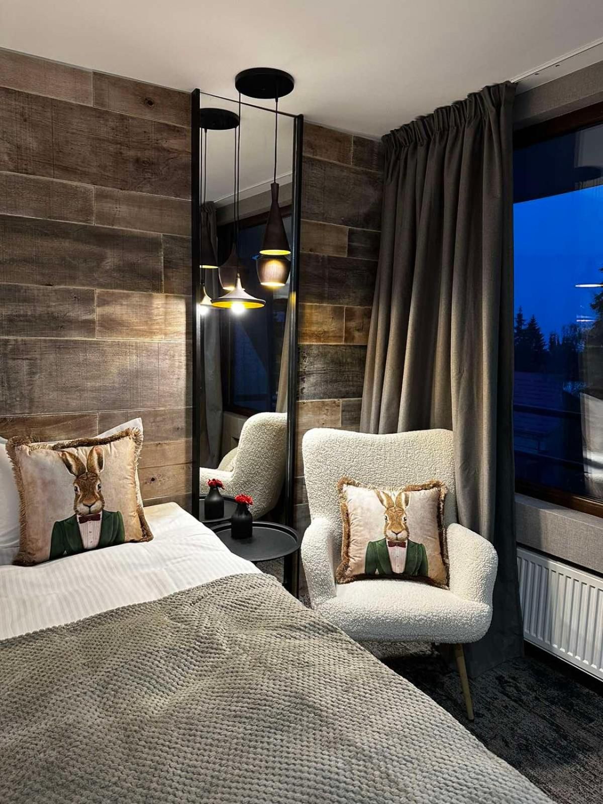 A modern hotel room with a large mirror, a plush armchair, a bed with decorative pillows, and a view of the outdoors.