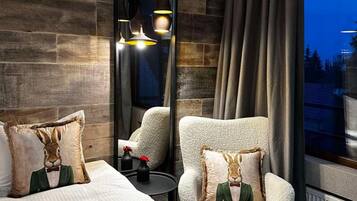 A modern hotel room with a large mirror, a plush armchair, a bed with decorative pillows, and a view of the outdoors.