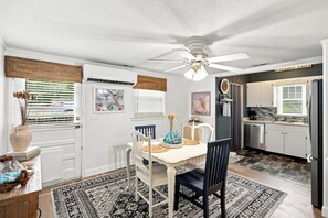A dining area with a white table, black chairs, a ceiling fan, and a kitchen with a refrigerator and stove. - Cozy Holden Beach dog friendly beach cottage. Only 1.4 miles to Holden Beach on mainland side. (Supply)
