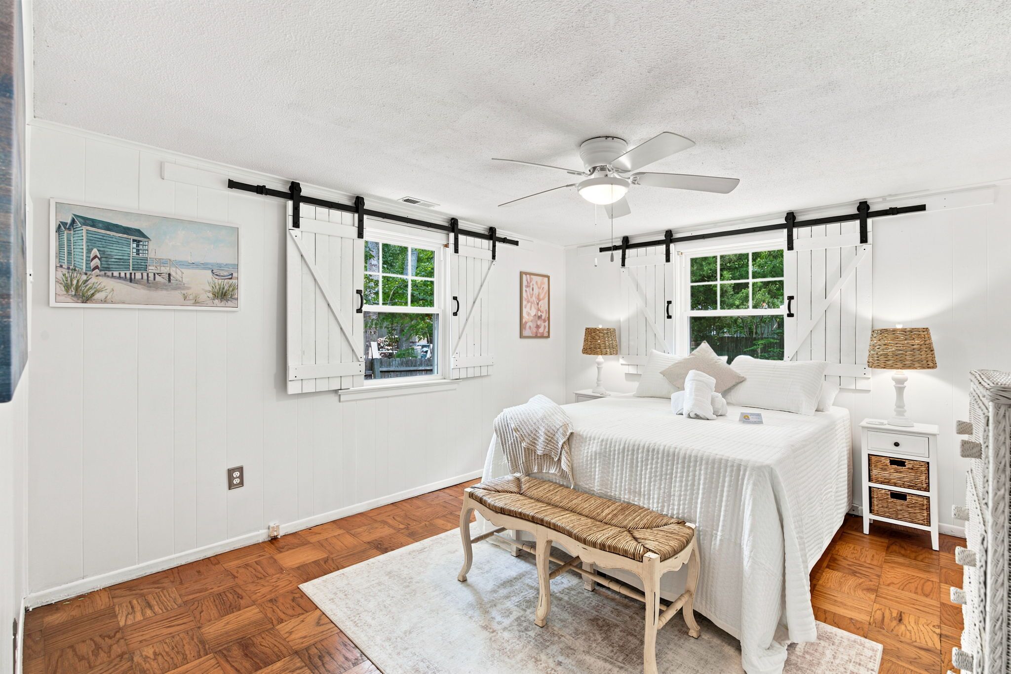 A bedroom with a bed, bedside tables, a bench, a ceiling fan, and two windows with shutters.